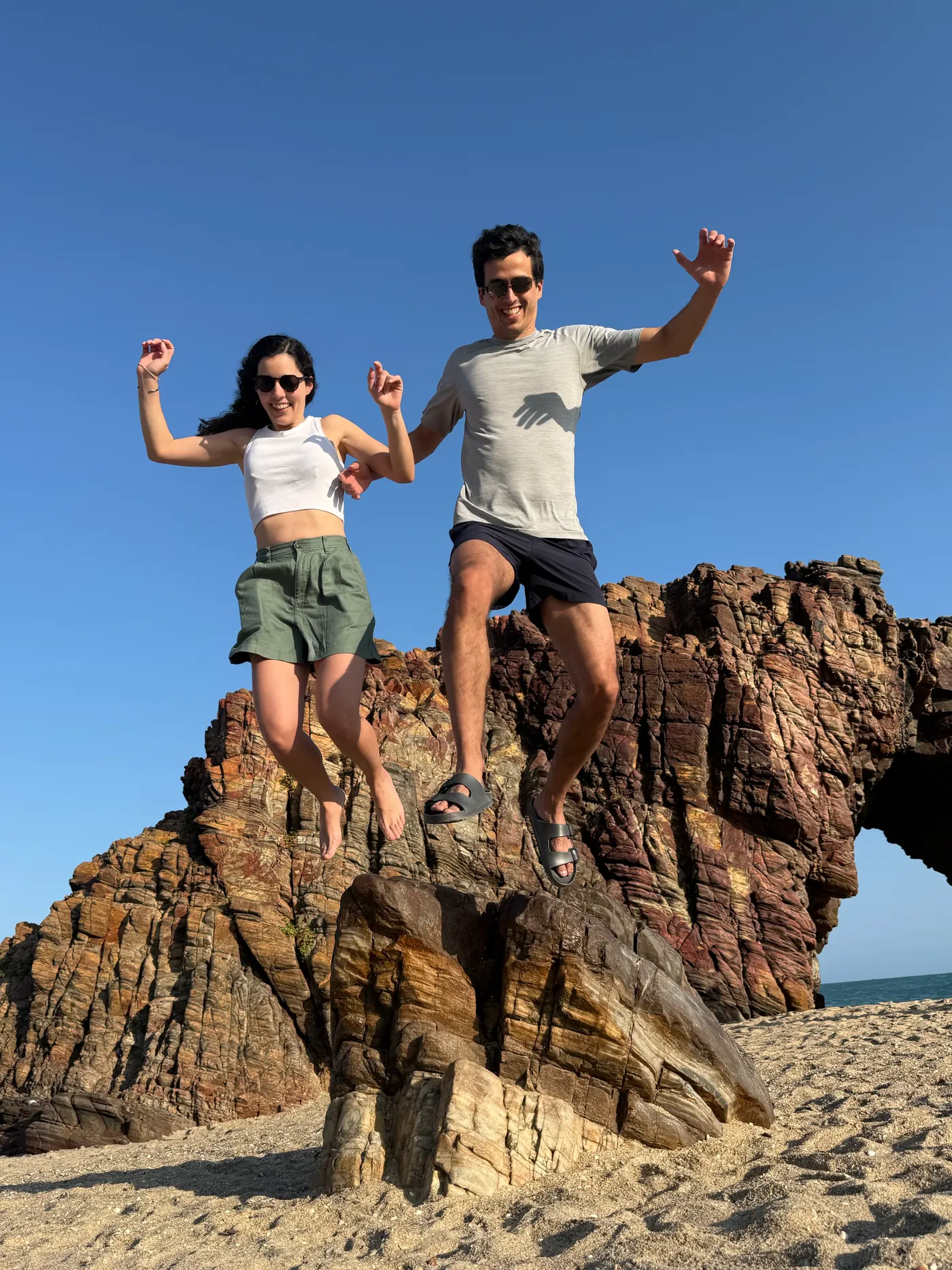 Vale y Fede saltando desde una roca en la playa, bajo un cielo azul.